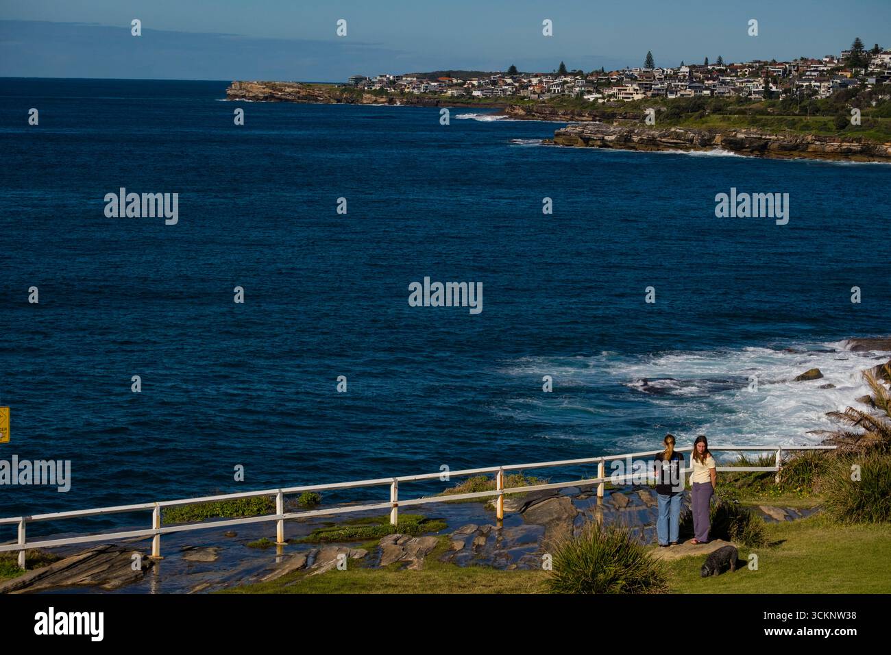 Malerischer Küstenpfad mit Blick auf ein riesiges blaues Meer mit weit entfernten Häusern und zwei Individuen, die die Natur genießen und natürliche Schönheit mit menschlichem Können verbinden Stockfoto