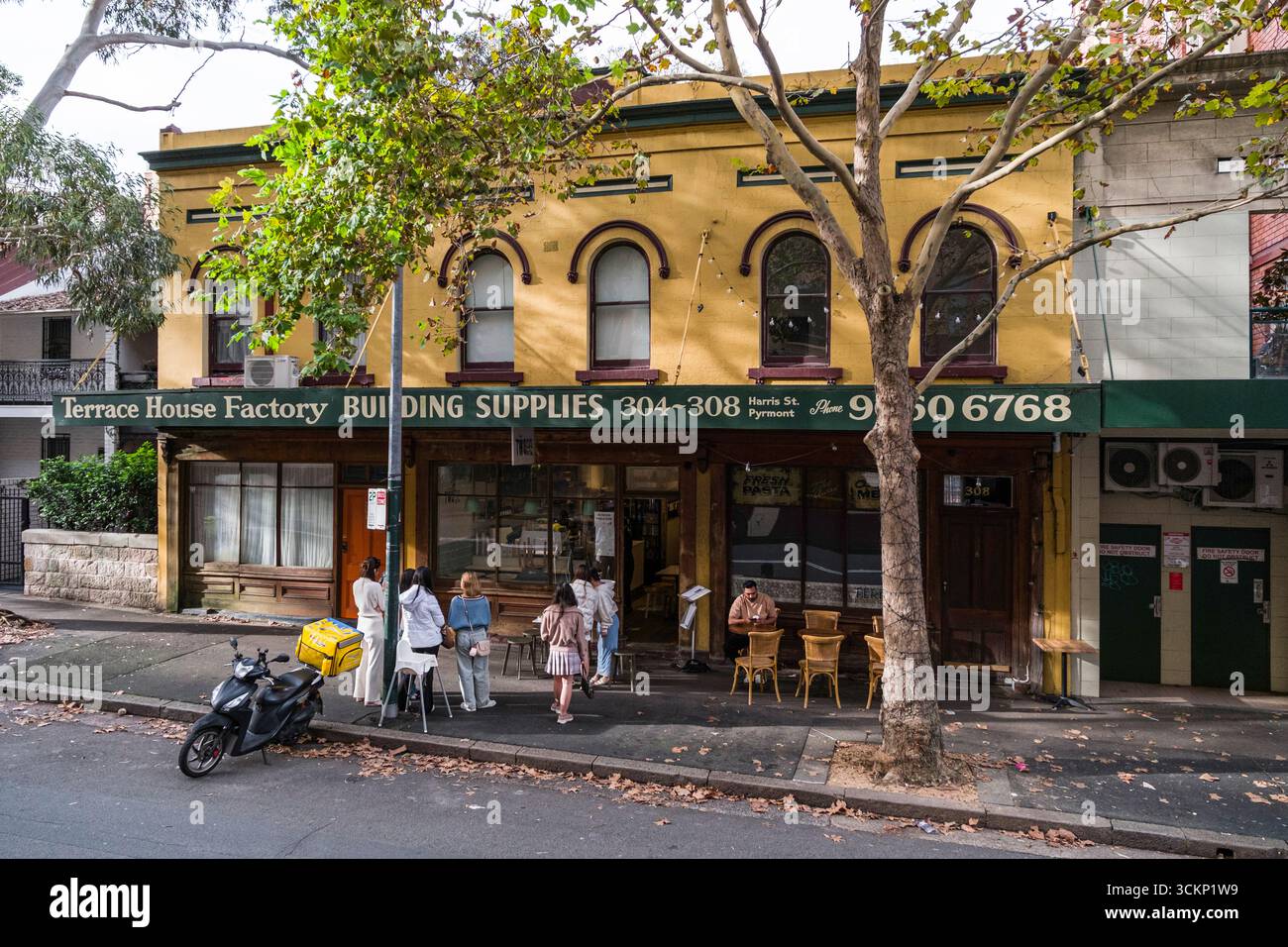 Ein markantes gelbes Gebäude mit Cafés auf Straßenebene mit Sitzgelegenheiten im Freien zwischen Stadtbäumen, die an eine lebhafte urbane Szene erinnern, Sydney Stockfoto