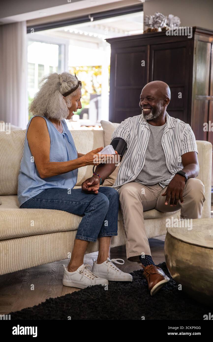 Verschiedene Seniorenpaare, die im Wohnzimmer auf einem Sofa sitzen und mit digitaler Blutdruckmanschette den Blutdruck messen Stockfoto