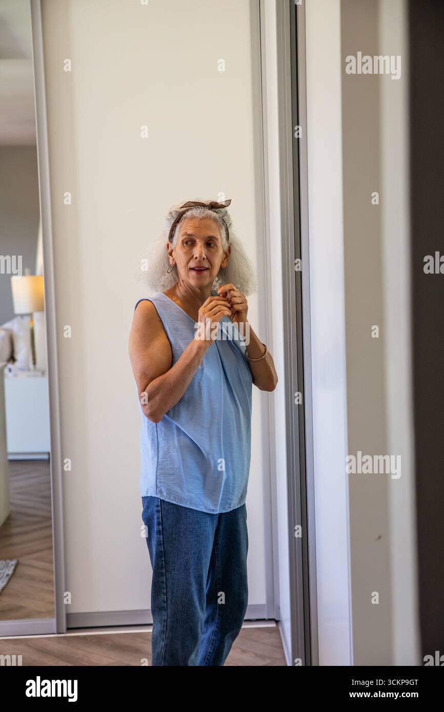 Weiße Seniorin, die im Ankleidebereich steht und Ohrringe in der Nähe der Spiegeltür anstellt Stockfoto