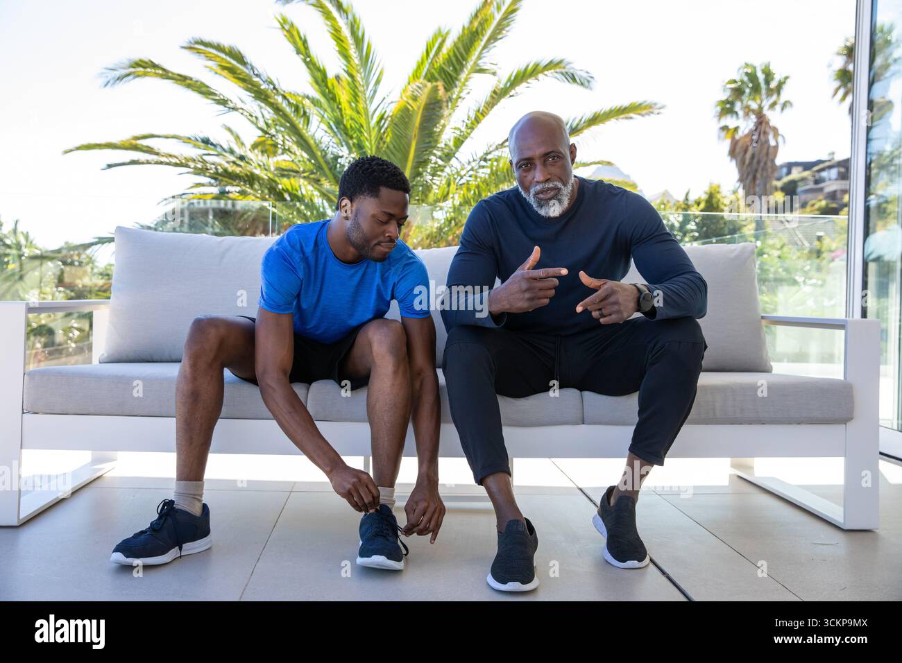 afroamerikanischer Vater, Sohn in Sportkleidung, der Schuhe bindet, plaudert auf der Terrasse in der Nähe von Glasgeländern Stockfoto