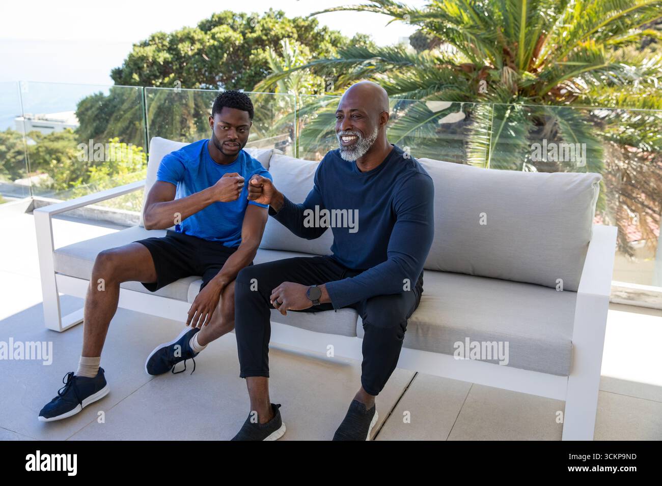afroamerikanischer Vater und Sohn in Sportkleidung prallt Fäuste auf Terrassensofa mit Glasgeländer Stockfoto
