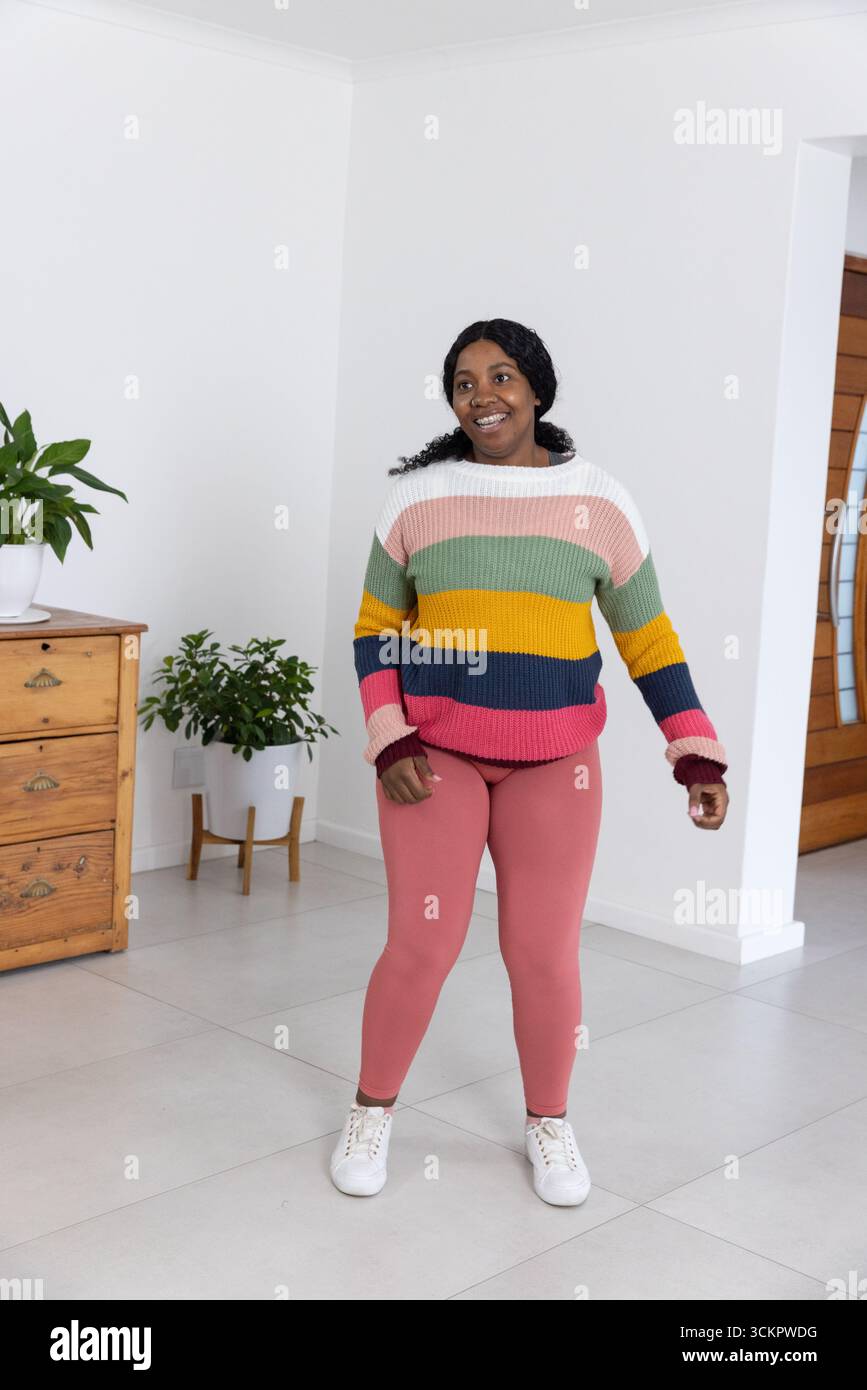 afroamerikanerin, die zu Hause im Pullover steht, Leggings in der Nähe von Schubladen, Pflanzen und Tür Stockfoto