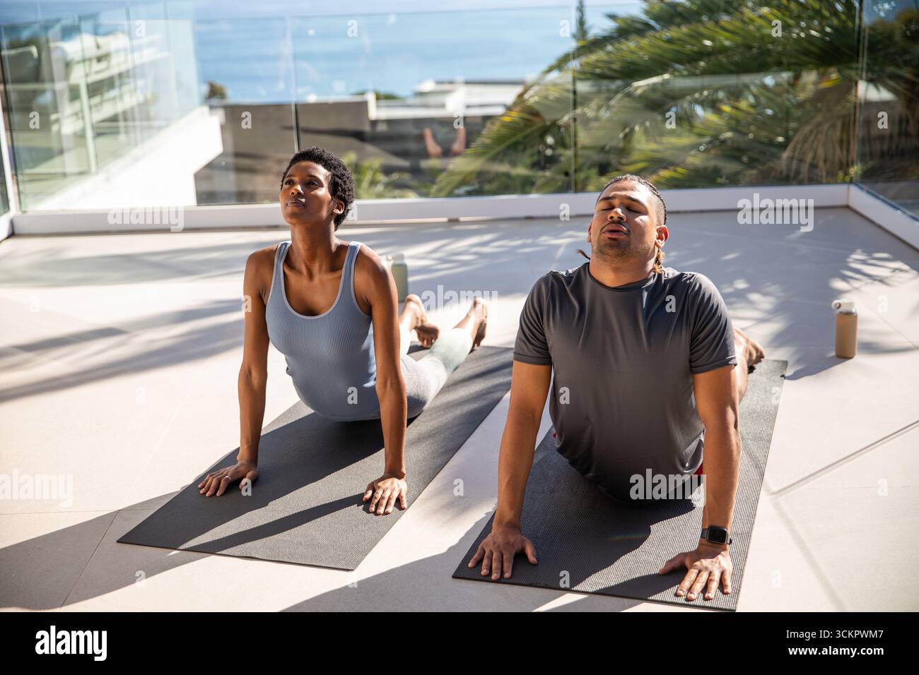 Paare üben auf Yoga-Matten auf der Terrasse mit Blick auf das Meer mit einer Wasserflasche einen nach oben gerichteten Hund Stockfoto