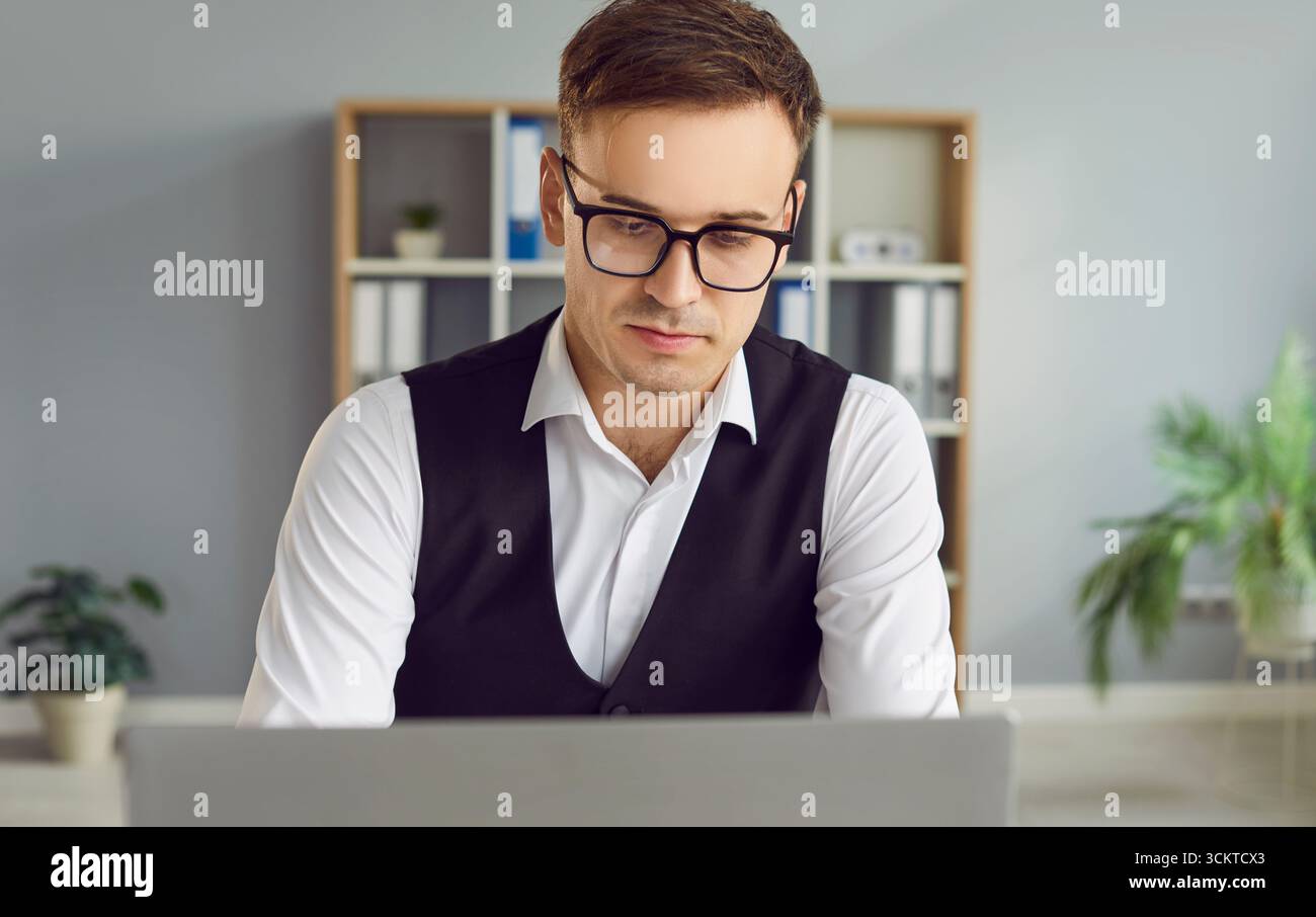 Ernsthafter junger Geschäftsmann mit Brille, der im Büro an einem Laptop arbeitet Stockfoto