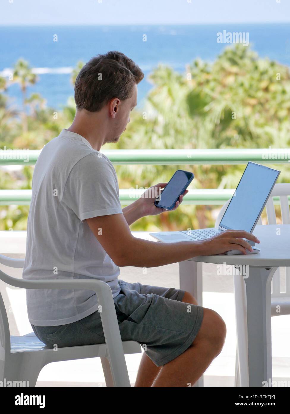 Junger Mann, der mit Laptop und Smartphone auf einem Balkon arbeitet. Konzept für den Lebensstil digitaler Nomaden, freiberufliche Arbeit, mobile Produktivität und Arbeit Stockfoto