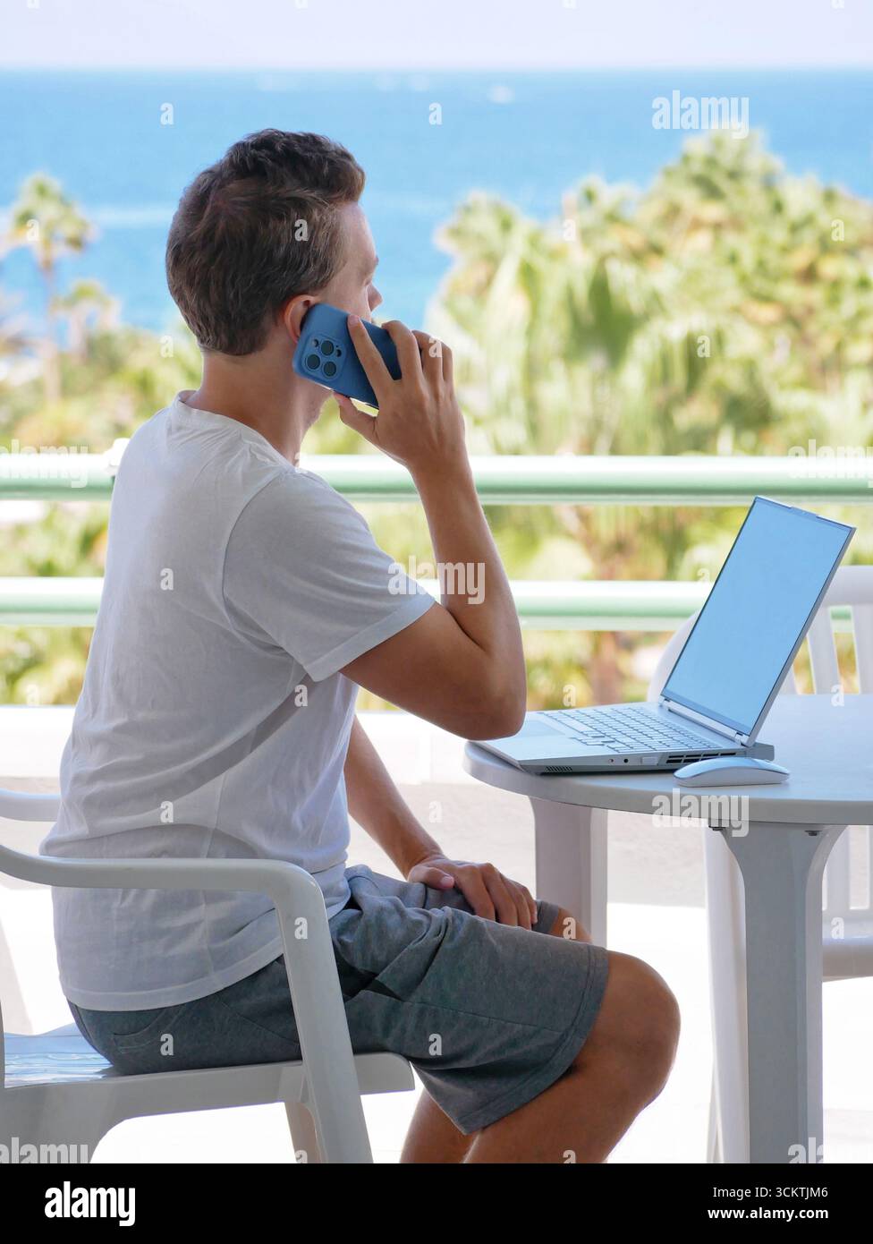 Mann, der ferngesteuert am Meer arbeitet, mit einem Smartphone spricht, während er draußen einen Laptop benutzt. Konzepte wie Remote-Arbeit, Arbeit und Standortunabhängigkeit Stockfoto