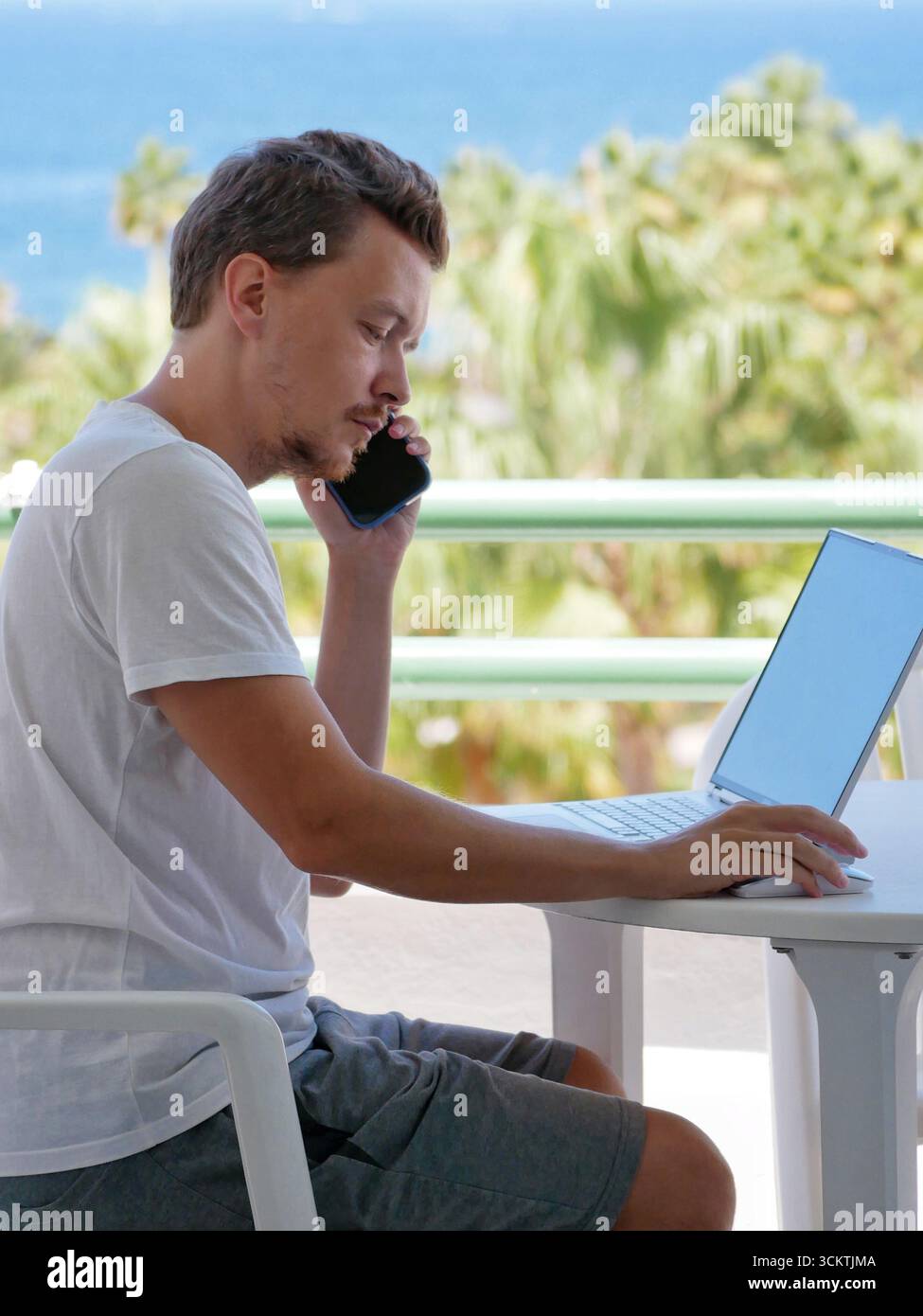 Mann, der ferngesteuert am Meer arbeitet, mit einem Smartphone spricht, während er draußen einen Laptop benutzt. Konzepte wie Remote-Arbeit, Arbeit und Standortunabhängigkeit Stockfoto