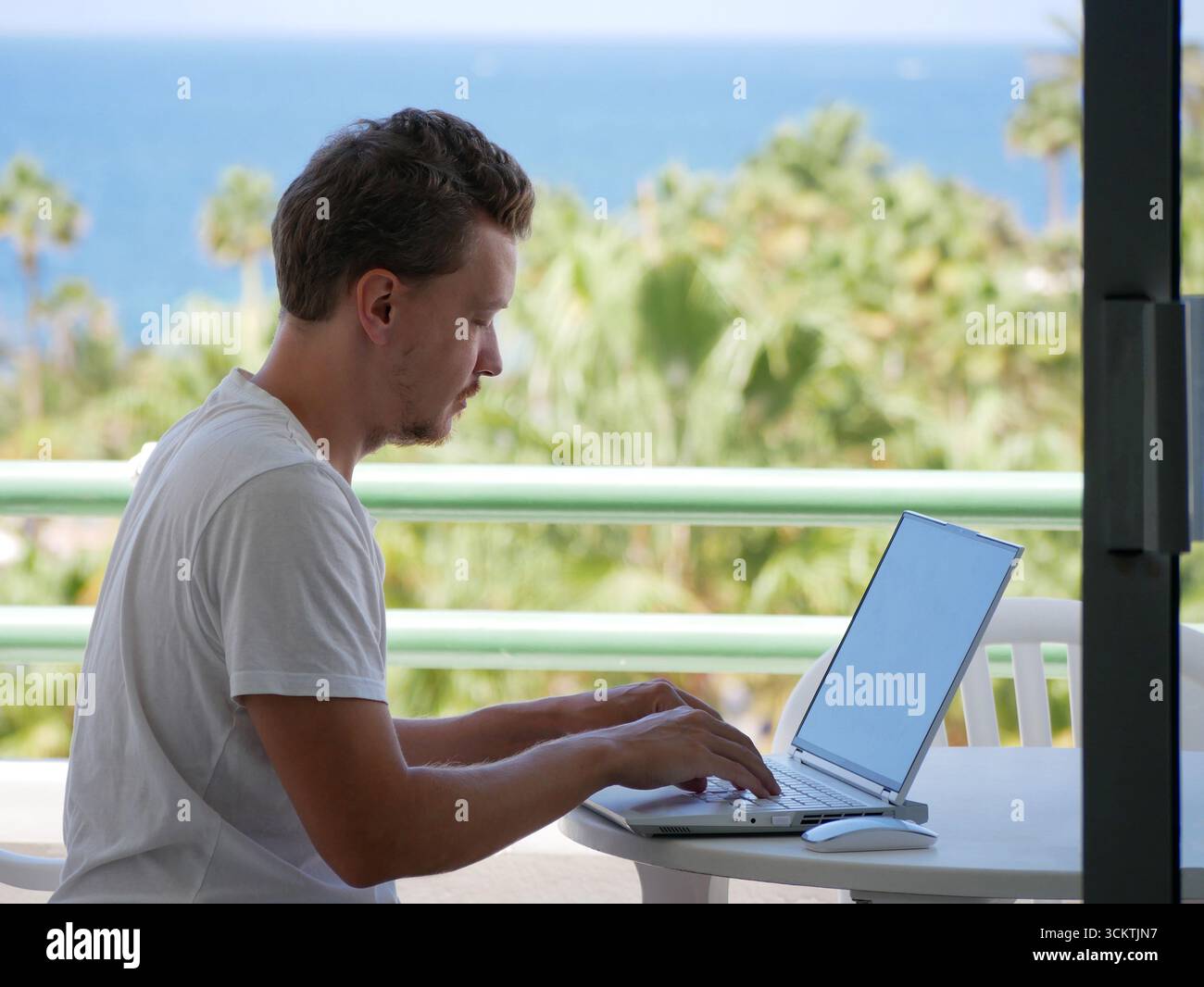 Ein junger Mann arbeitet ferngesteuert an einem Laptop, während er draußen an einem tropischen Ort sitzt. Themen wie freiberufliche Arbeit, Lebensstil digitaler Nomaden, Work-Life balan Stockfoto