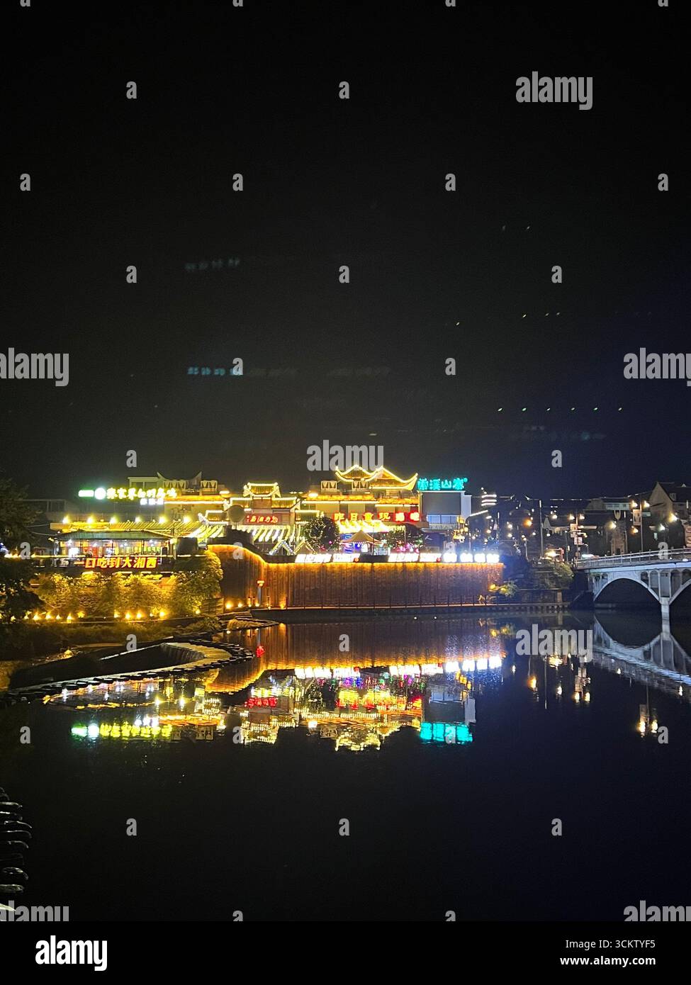 Zhangjiajie bei Nacht – Neonreflexionen tanzen auf dem Fluss unter den leuchtenden Lichtern der Stadt. Stockfoto