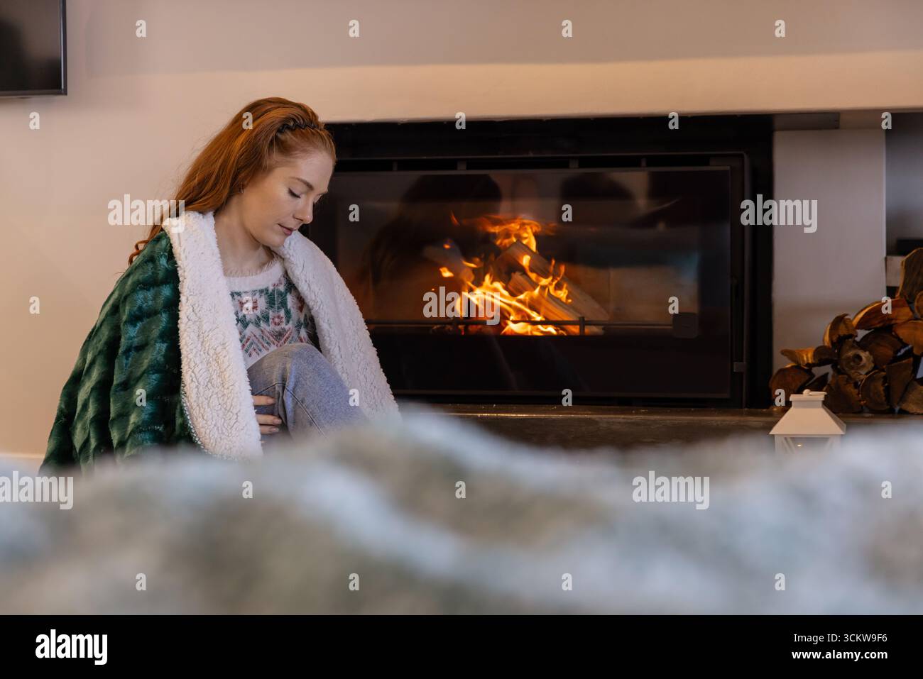 Rothaarige Frau, die auf dem Boden bei einem Glaskamin im Wohnzimmer in einer Plüschdecke sitzt, Kopierraum Stockfoto