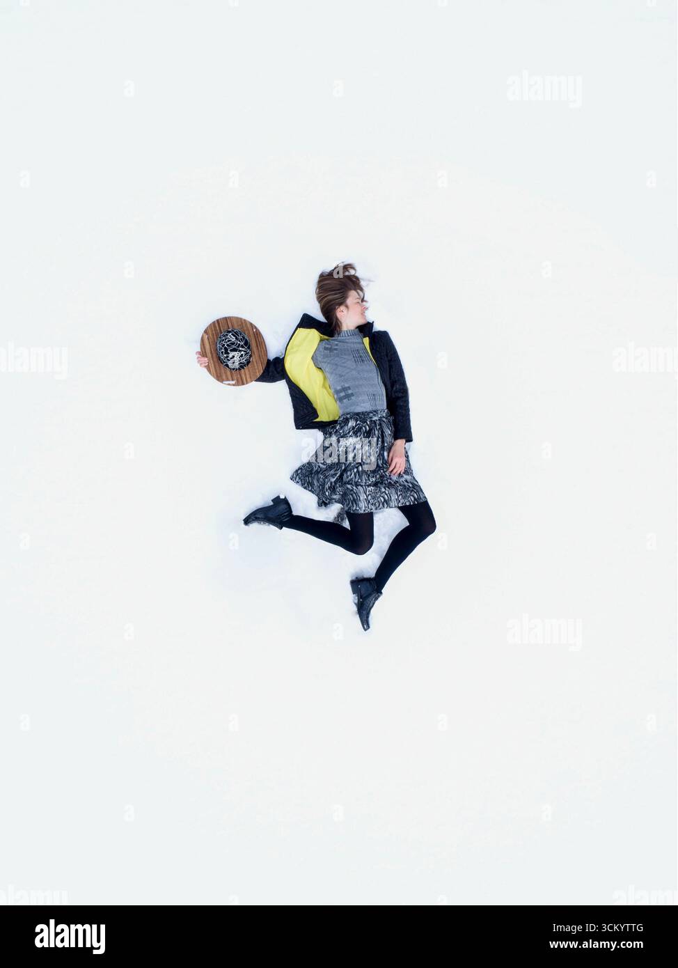 Schöne junge Frau, die auf weißem Schneeteppich neben dem Hut liegt. Oben erhobene Drohnenansicht des Modemodells, das im weißen Schnee bezahlt und entspannt. Stockfoto