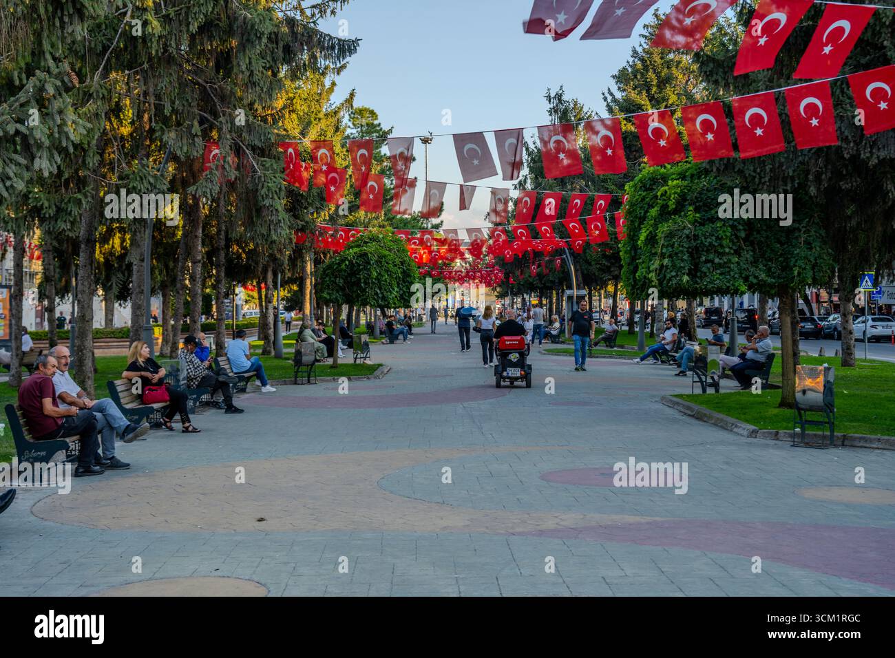 Die Leute sitzen auf Bänken und laufen entlang eines Parkgangs, der mit türkischen Fahnen geschmückt ist. Adapazarı, Sakarya, Türkiye – 09. September 2025. Stockfoto