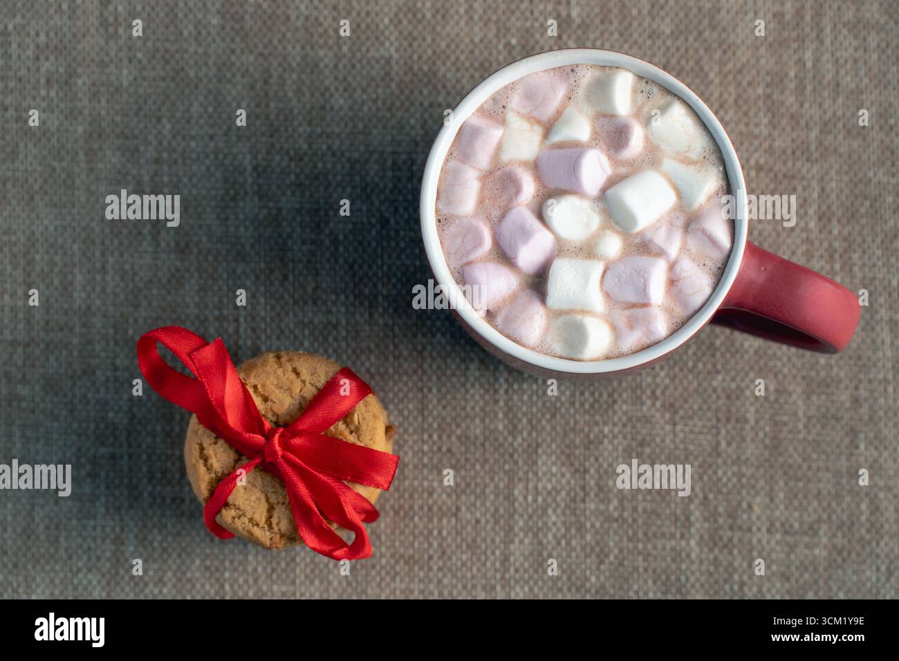Ein Blick von oben auf einen einzelnen Weihnachtskeks, der mit einem roten Band verbunden ist, und eine rote Tasse heiße Schokolade mit Marshmallows Stockfoto