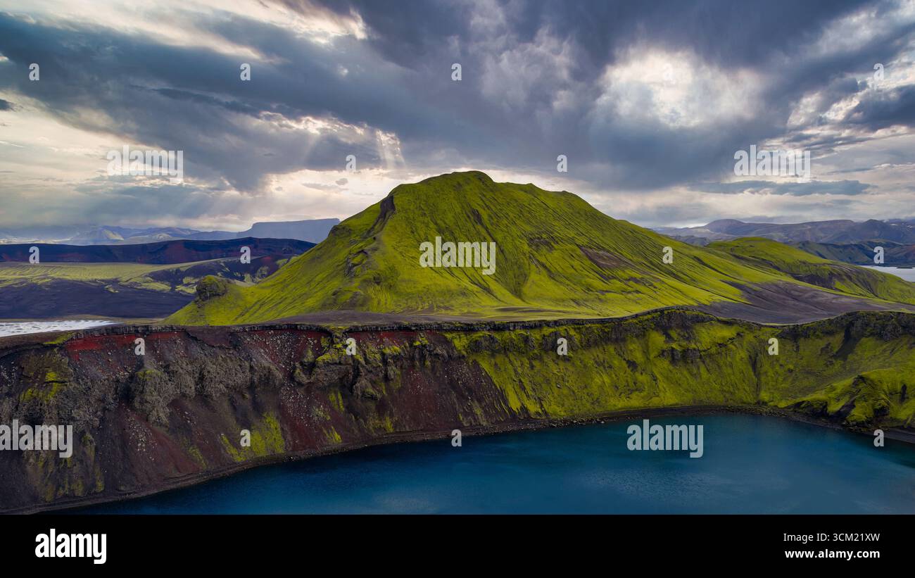 Aus der Vogelperspektive auf einen mit grünem Moos bedeckten vulkanischen Berg in Island mit einem tiefen blauen Kratersee, umgeben von rauem Gelände und einem wolkengefüllten Himmel. Stockfoto