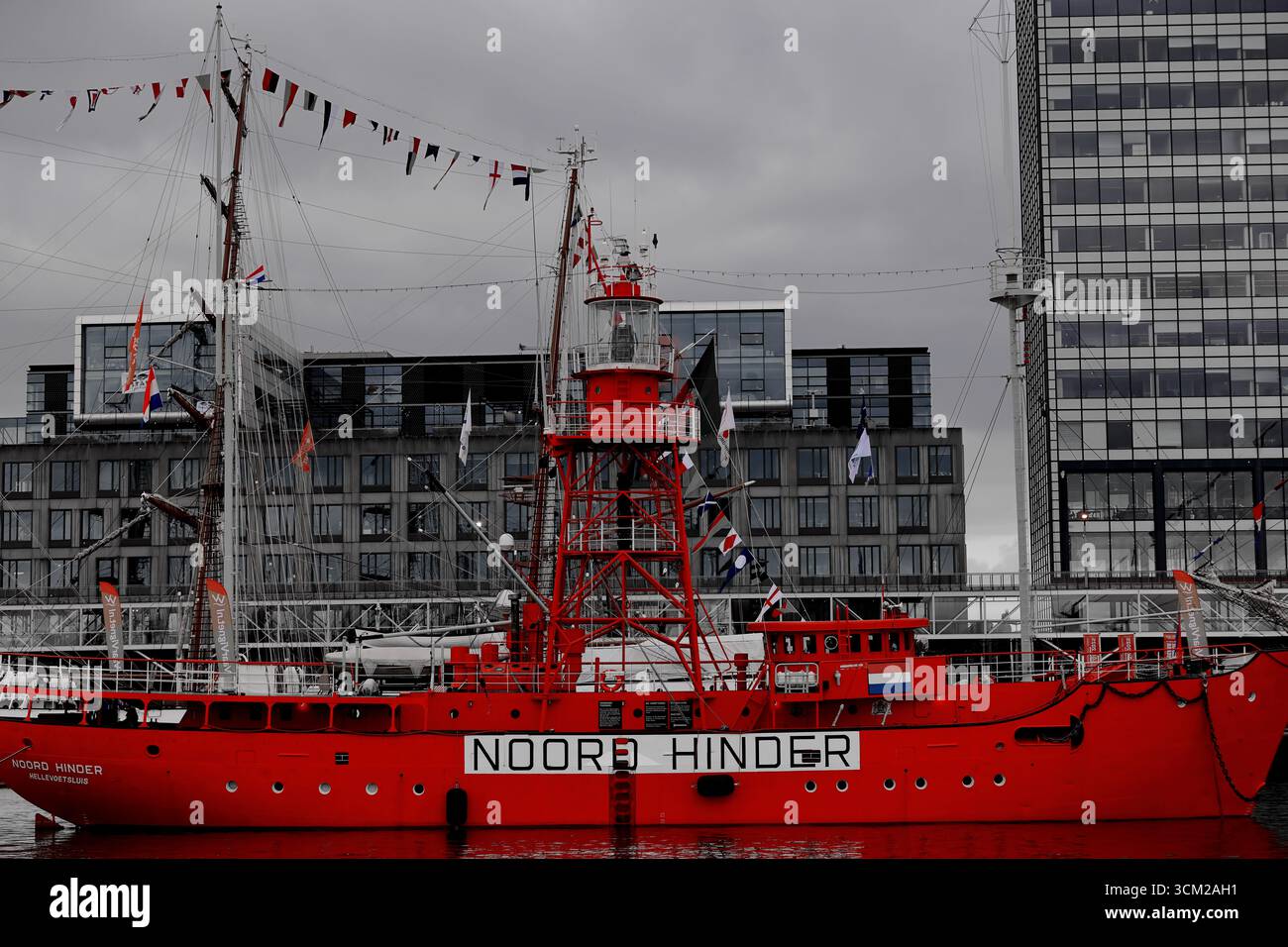 Lichtschip Noord hindern neben bei Sail 2025 in Amsterdam, Niederlande, das volle Rumpfprofil des roten Leuchtschiffes mit Fahnen und Stadtgebäuden dahinter. Stockfoto