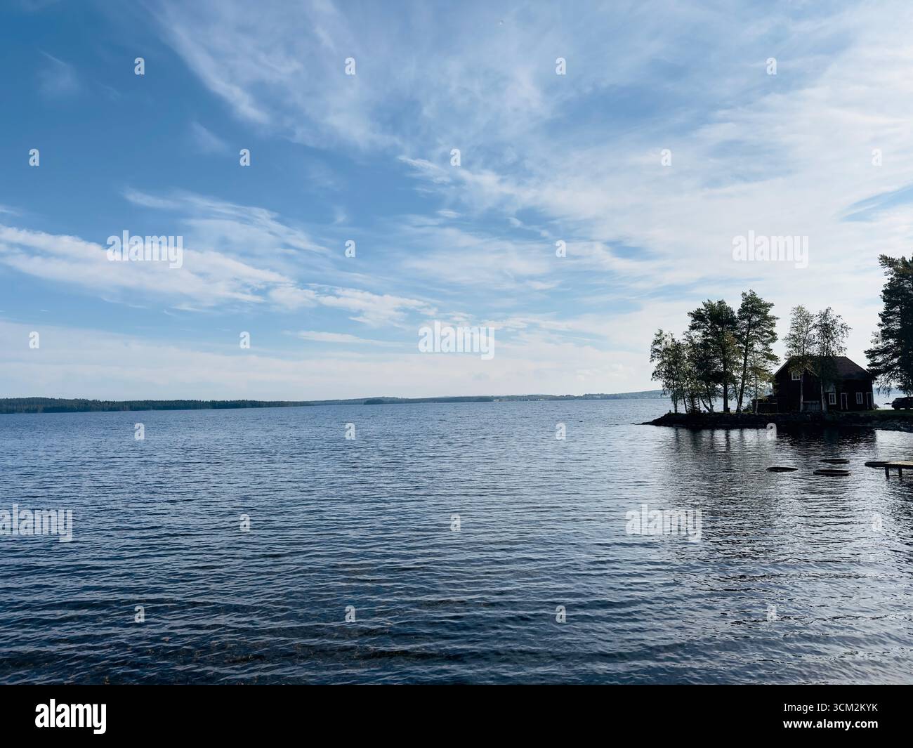 Malerischer Blick auf einen See mit einer kleinen Insel und einem Haus unter einem hellblauen Himmel. Stockfoto