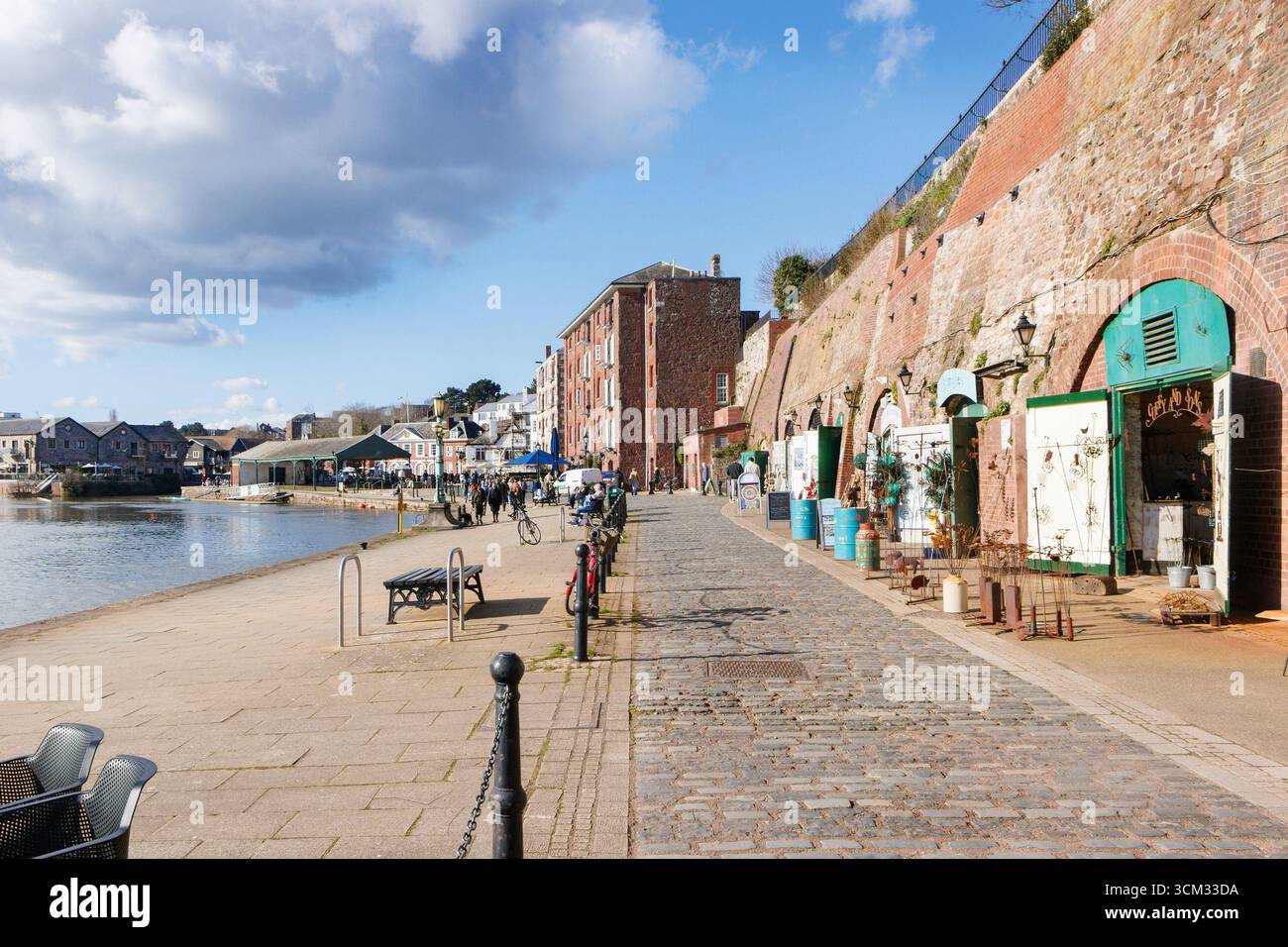 Malerischer Blick auf den Fluss exe auf den Quay in exeter devon mit historischen Steinhäusern, Geschäften und Cafés entlang einer gepflasterten Uferpromenade unter blauem Himmel Stockfoto