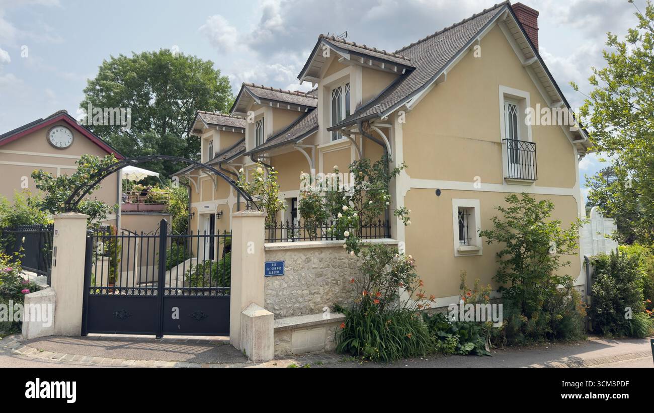 Ein cremefarbenes Haus mit einem dunklen Dach und schwarz akzentuierten Fenstern und Türen, umgeben von einem schwarzen Metalltor und üppigem Grün, unter einem teilweise bewölkten s Stockfoto