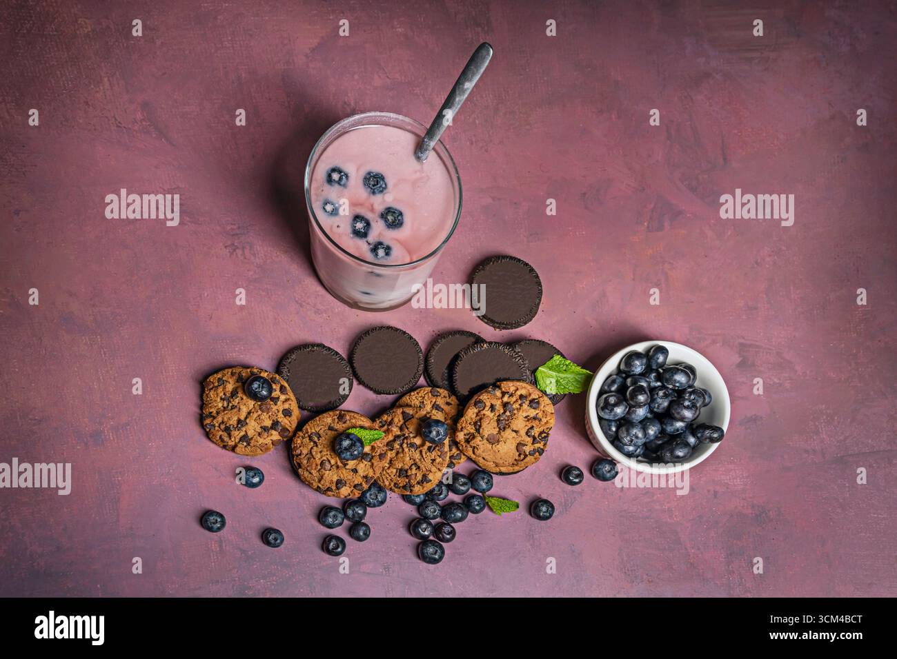Stillleben mit verschiedenen Keksen in verschiedenen Formen und Größen, darunter einige mit Heidelbeerbrocken, begleitet von einem erfrischenden Smoothie auf einem Licht, Stockfoto