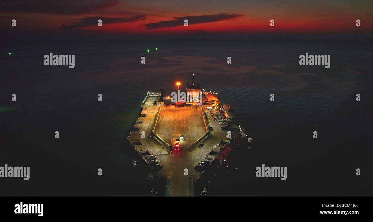 Luftdrohne einer Fähre, die nachts im Moo Koh Ang Thong National Marine Park in Thailand anlegt, mit Autos, die vom Boot fahren und einem dramatischen Sonnenuntergang im Hintergrund Stockfoto