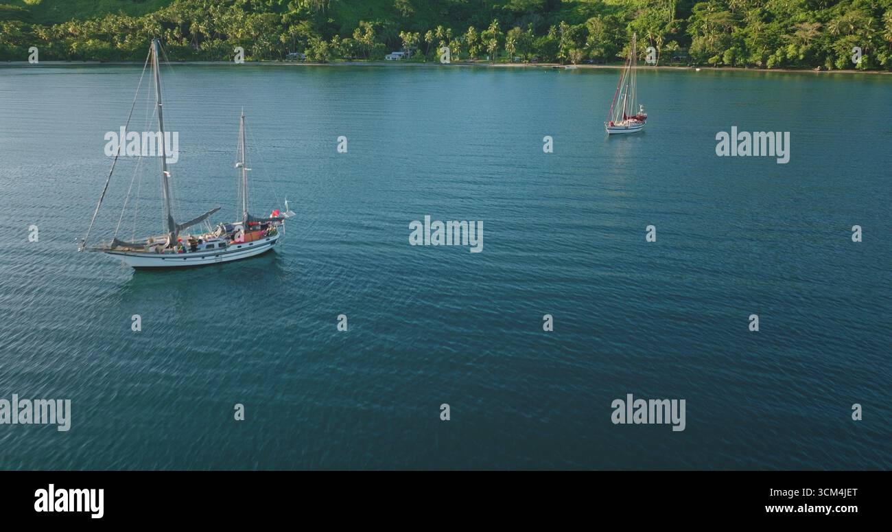 Fidschi-Inseln: Zwei Segelboote, die auf ruhigem blauem Wasser in der Nähe einer tropischen Küste schwimmen, üppige grüne Vegetation, die von einem warmen Sonnenuntergang beleuchtet wird. Wilde Naturreiselandschaft. Drohnenflug aus der Luft Stockfoto