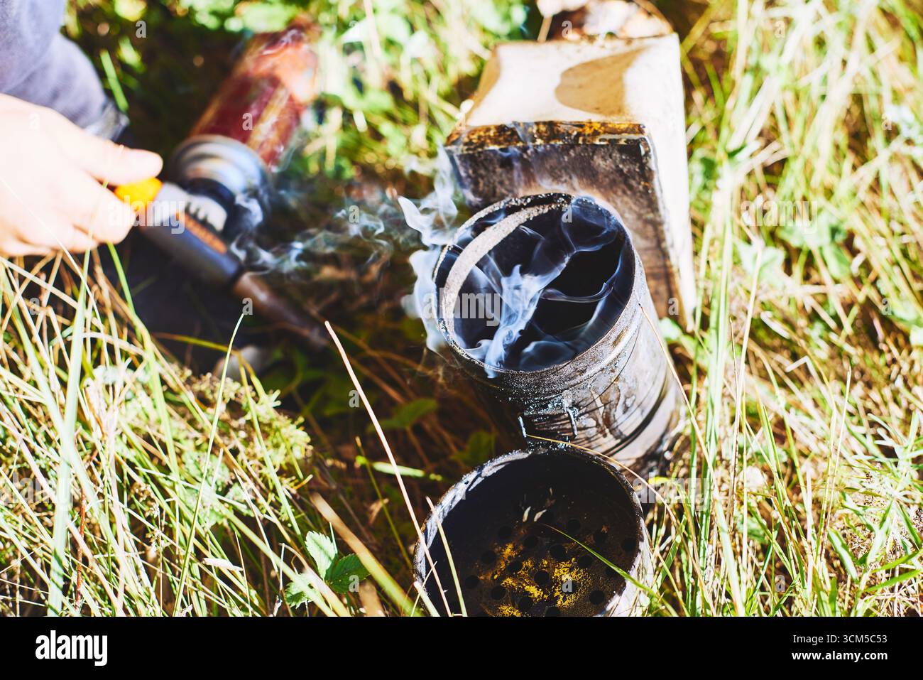 Handbetriebener Metallbienenraucher mit sichtbarem Rauch, der im Freien in grasbewachsenen Bereichen aufsteigt und sich auf die Imkerei vorbereitet, Ausrüstung und Werkzeuge auf dem Boden verstreut Stockfoto