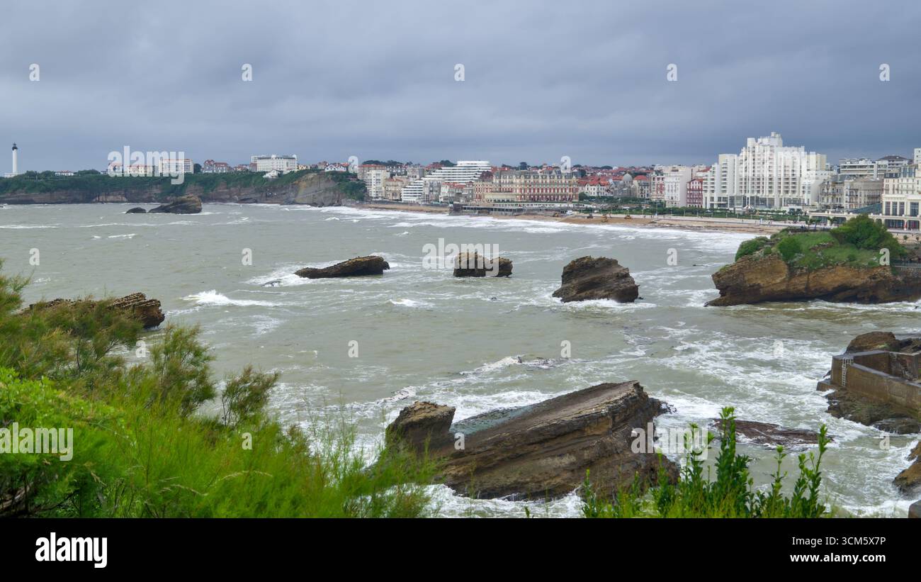 Küstenstraße in Biarritz in der Nähe des Rocher de la Vierge mit Wellen des Atlantiks und Stadtblick bei rauem Wetter. Stockfoto
