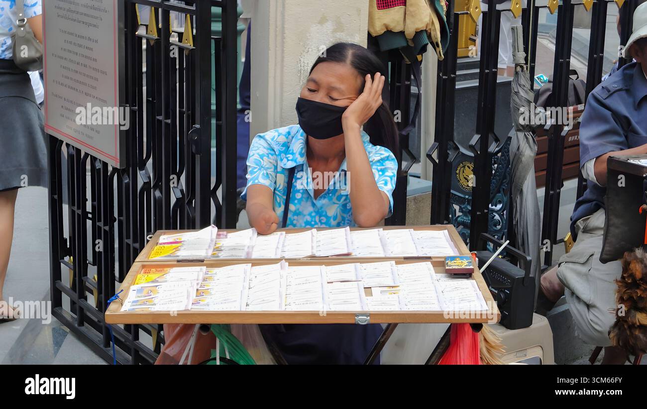 Eine Frau, die an einem kleinen Tisch sitzt, mit dem Kopf auf der Hand und Lottoscheine verkauft. Stockfoto