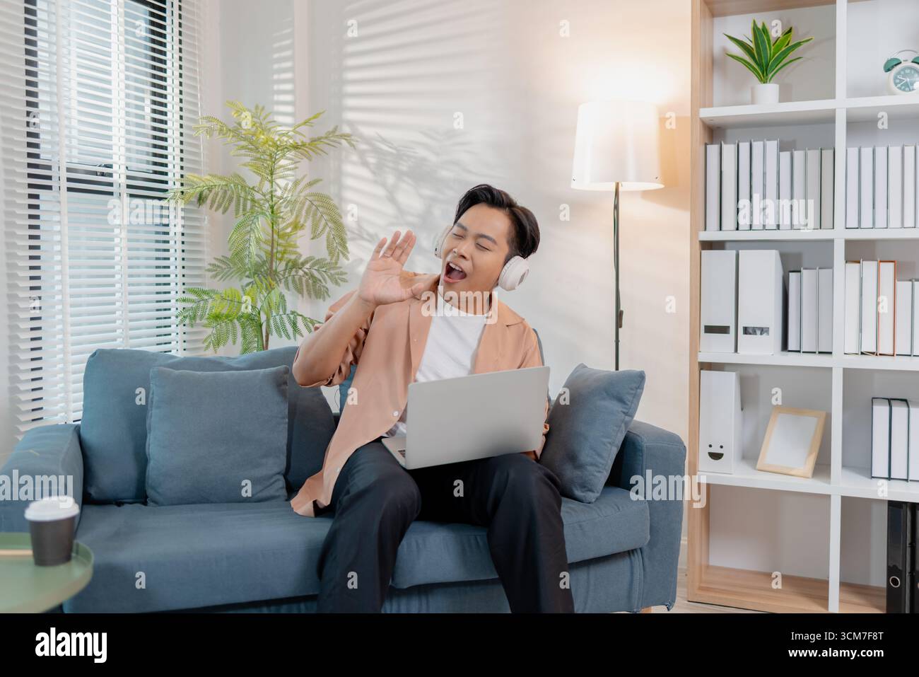 Ein fröhlicher junger Mann sitzt auf einem Sofa und singt mit Leidenschaft, während er einen Laptop hält. Seine fröhlichen Gesten und sein heller Ausdruck spiegeln den Spaß der Musik wider Stockfoto
