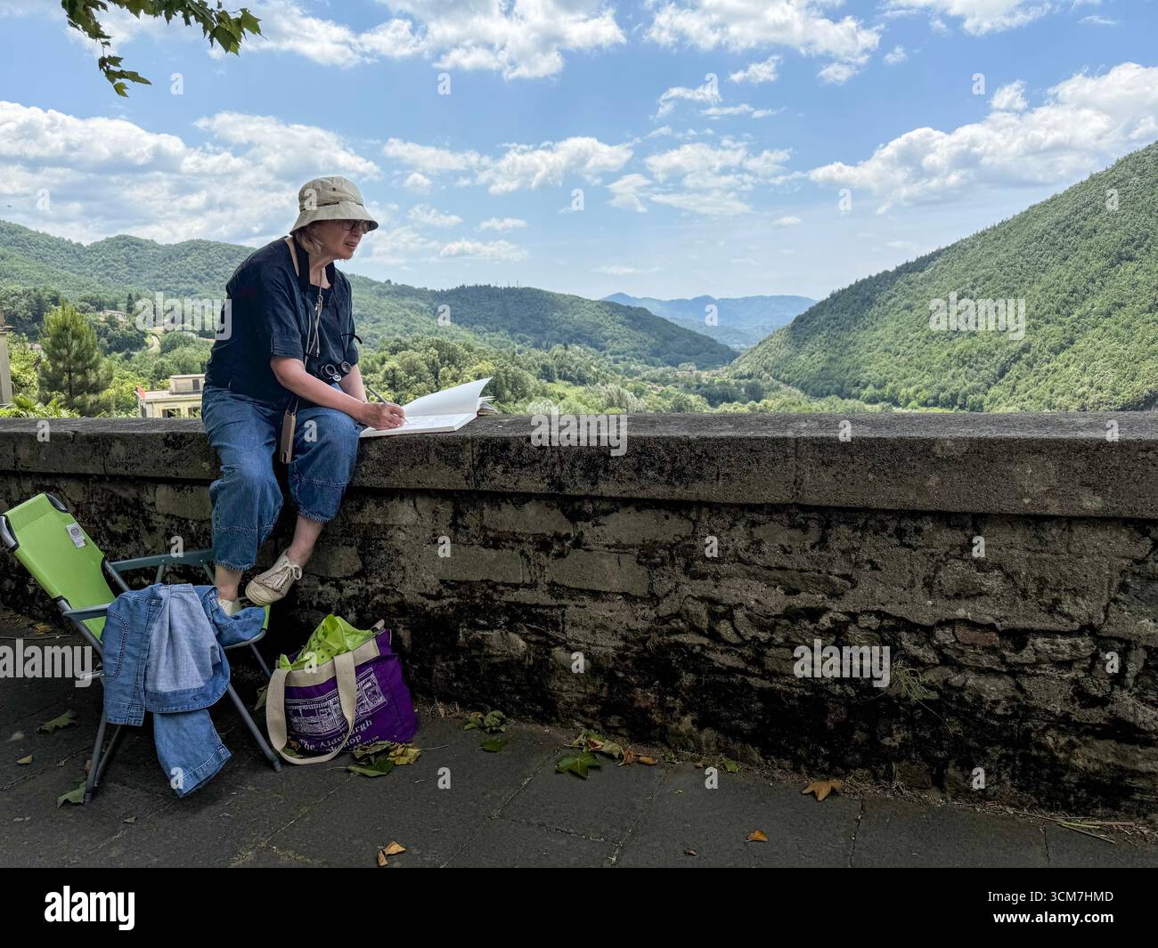 Eine Künstlerin, die auf einer Wand sitzt und eine Szene der steilen bewaldeten Berghänge und auf dem Talboden des Valle Ombrosa (Shady Valley) wi malte Stockfoto