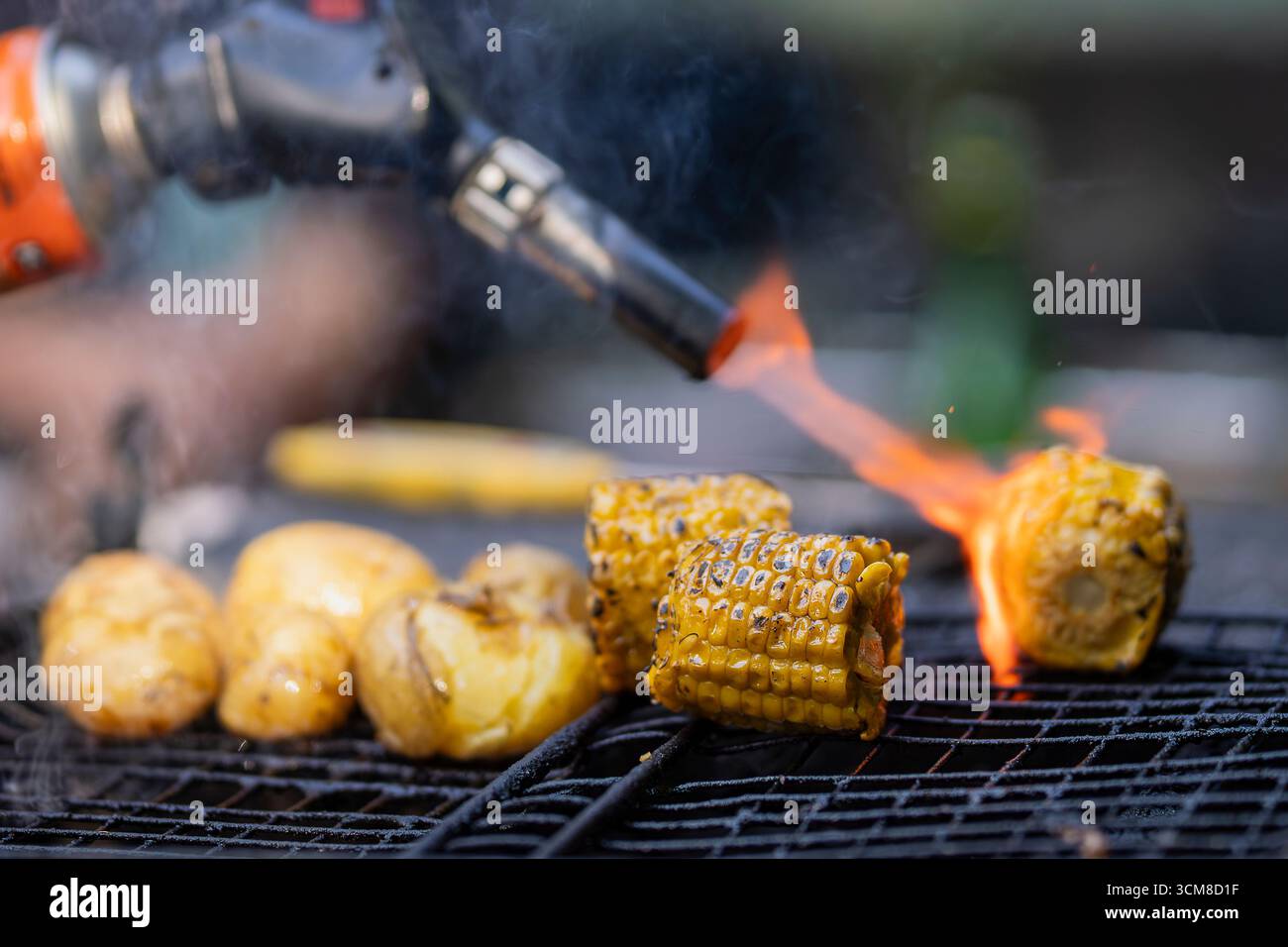 Auf heißem Kohlefeuer gegrillter Zuckermais. Von der Seite gesehen. Reife goldene Maiskolben und Kartoffeln auf dem Grill mit Rauch und Flamme. Vegetarisches Barbecue über dem f Stockfoto