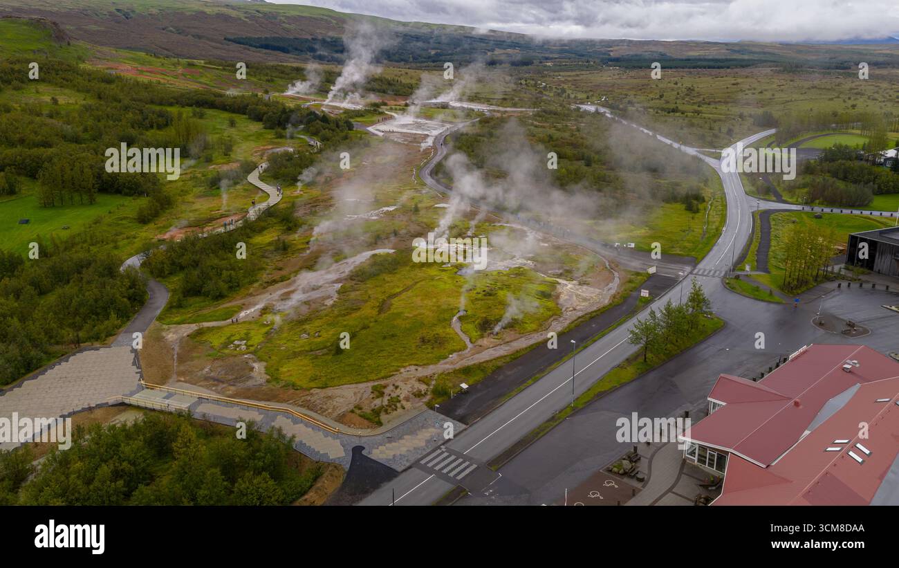 Luftbild von Haukadalur in Island mit dampfenden Lüftungsöffnungen, sprudelnden heißen Quellen, gewundenen Wegen, üppigem Grün und einem rot überdachten Gebäude. Stockfoto