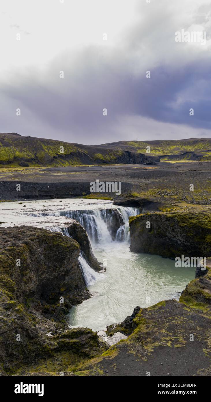 Ein kleiner Wasserfall fließt in einen türkisfarbenen Pool, umgeben von vulkanischen Klippen und grünem Moos. Die Luftaufnahme fängt das zerklüftete isländische Gelände ein. Stockfoto