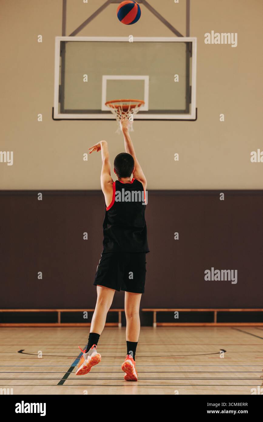 Ein junger Basketballspieler in sportlicher Kleidung übt seinen Schuss auf einem Hallenplatz. Den Moment des Loslassens des Balls festhalten und die Präzision hervorheben Stockfoto