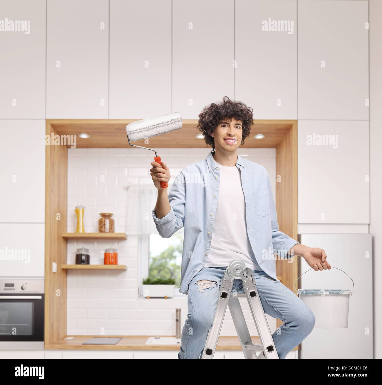 Junger Mann, der auf einer Leiter sitzt, mit Malerwalze und Eimer in einer Küche Stockfoto