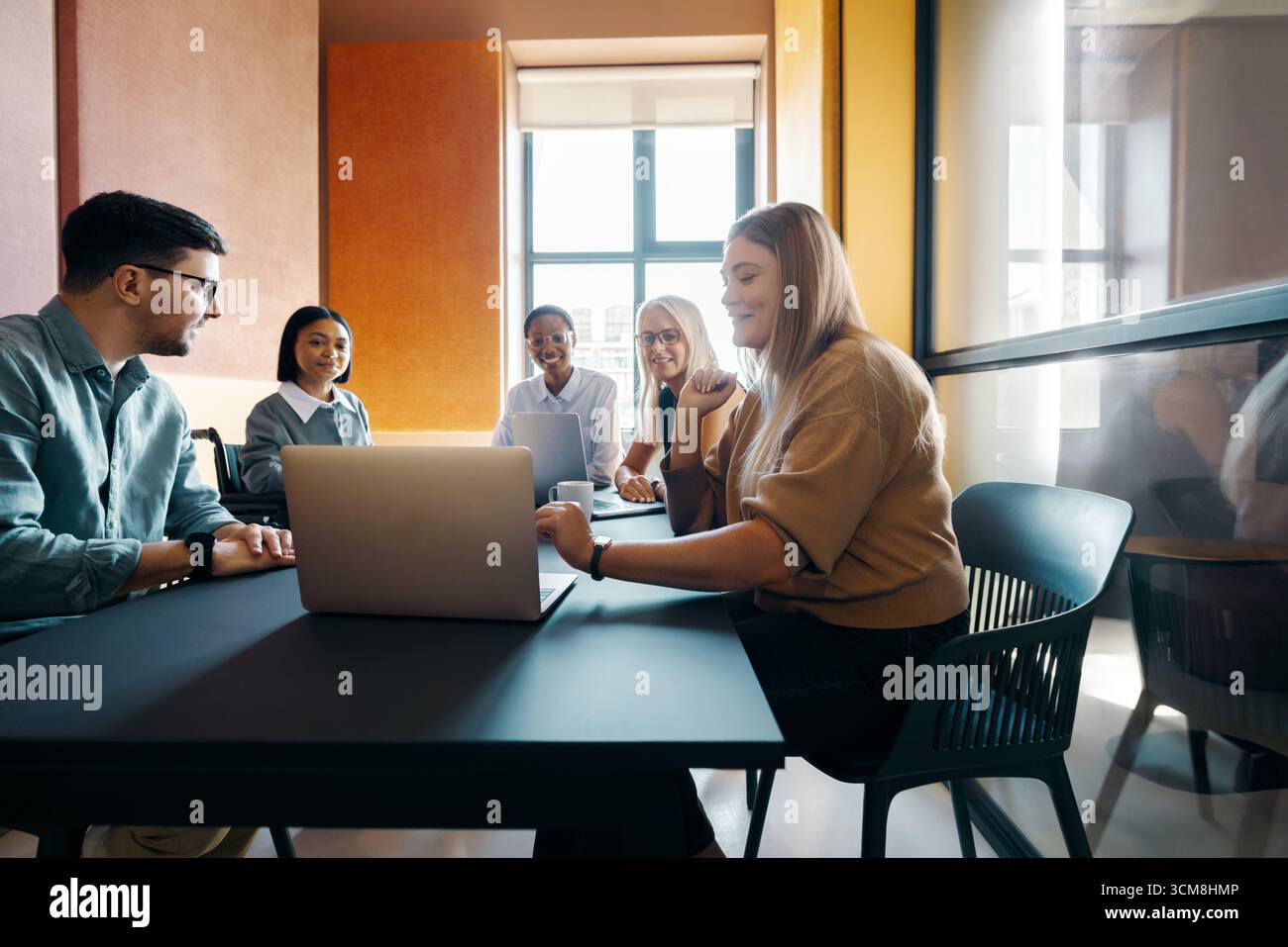 Gruppe junger Berufstätiger, Männer und Frauen, die an einem gemeinsamen Tisch in einer hell beleuchteten, modernen Büroumgebung arbeiten. Sie teilen die ID Stockfoto