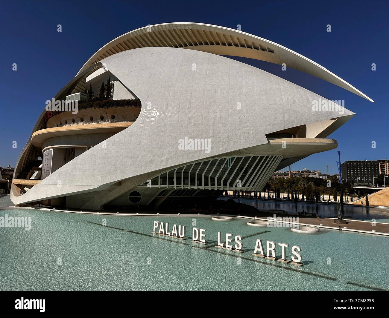 Das Bild zeigt den spektakulär gestalteten Palau de les Arts Reina SofÜa in Valencia, Spanien, ein Opernhaus mit markanter, futuristischer Architektur und dynamischen Formen. Das Gebäude ist von einem flachen, türkisfarbenen Wasserbecken umgeben, in dem sich die Schrift „Palau de les Arts“ widerspiegelt. Die Szene vermittelt Innovation, urbane Eleganz und die ausdrucksstarke Technik eines der architektonischen Wahrzeichen Valencias. Stockfoto