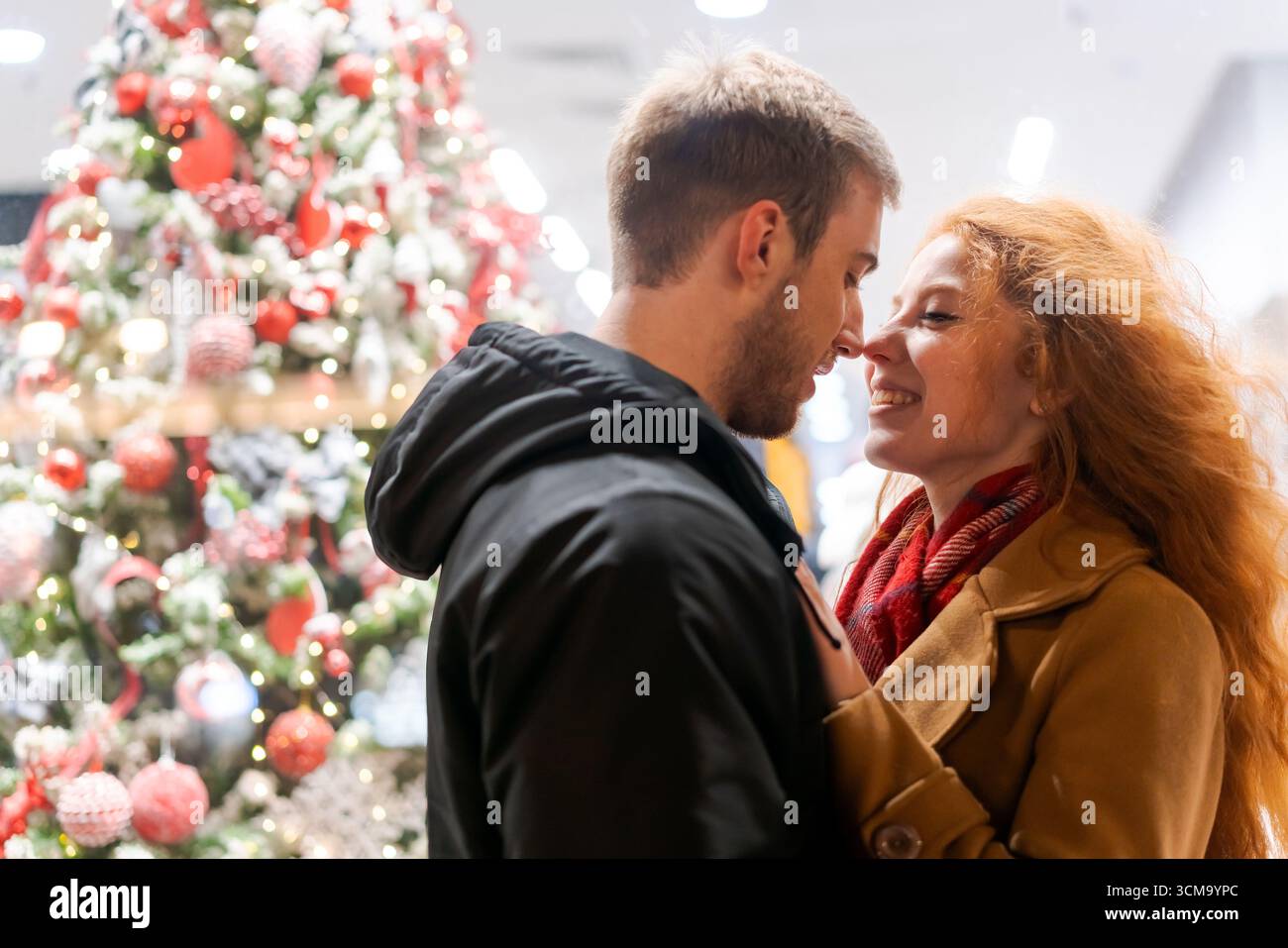Mann und Frau umarmen sich vor einem Weihnachtsbaum. Der Mann trägt eine schwarze Jacke und die Frau einen roten Schal Stockfoto