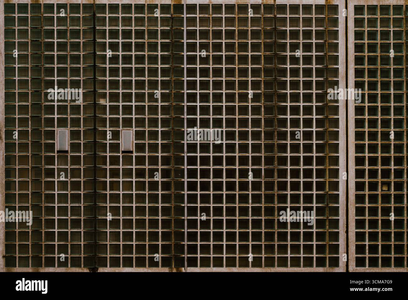 Geometrische Serenity: Milchglasfassade diese alte Milchglasfassade in Maastricht präsentiert ein ruhiges, aber auffälliges geometrisches Muster, bestehend aus unzähligen Quadraten, die ein nahtloses Gitter über die Oberfläche der Struktur bilden. Jedes Quadrat fängt das Licht anders ein und erzeugt ein subtiles Mosaik aus Farbtönen, die sich mit dem wechselnden Tageslicht verändern. Stockfoto