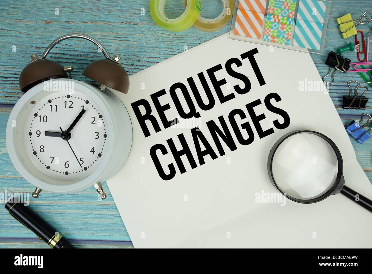 Anfrage ändert Text auf einem Papier, umgeben von Bürobedarf, einer Lupe und einer Uhr auf einer blauen Holzoberfläche Stockfoto