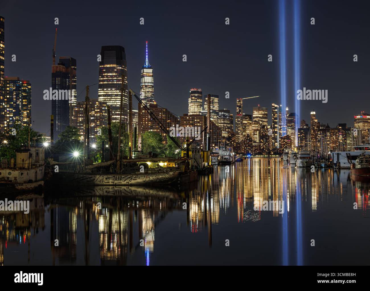 Nächtlicher Blick auf die Skyline von New York City mit den Tribute-in-Light-Gedenkstrahlen, die in den Himmel strahlen und sich im Hudson River neben beleuchtet spiegeln Stockfoto