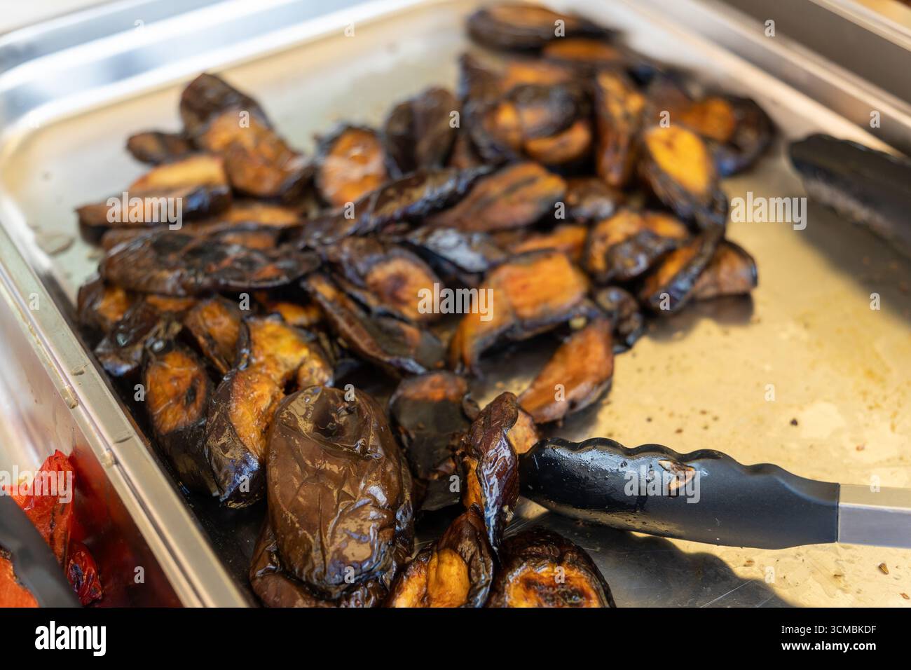 Gebackene Auberginen auf einem Tablett bei einem Grillbuffet. Serviert mit Zangen zum Selbstbedienung. Stockfoto