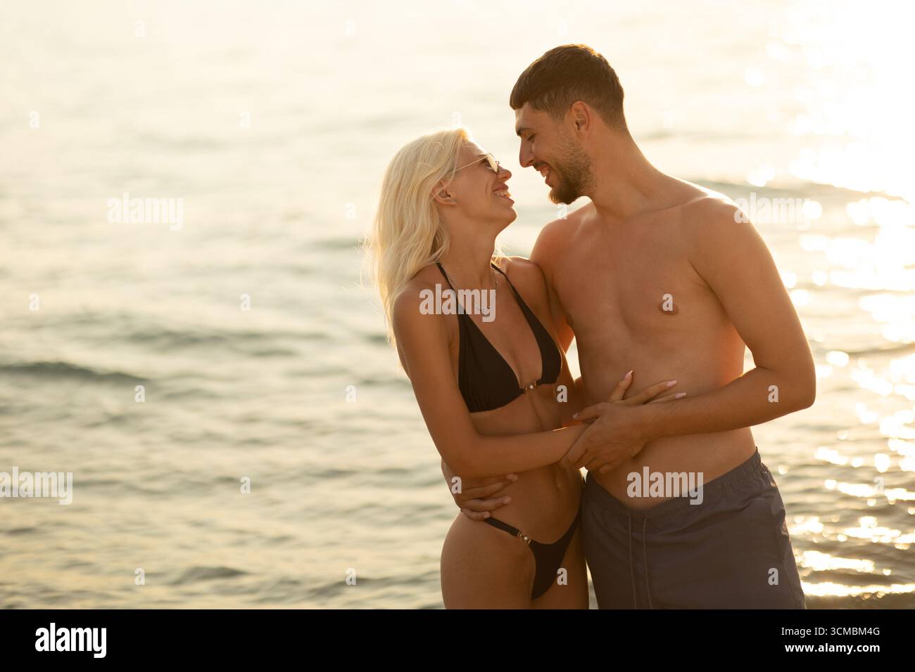 Zwei Personen teilen sich eine fröhliche Umarmung am Strand, während sie sich im warmen Glanz des Sonnenuntergangs über dem ruhigen Meer erfreuen. Stockfoto