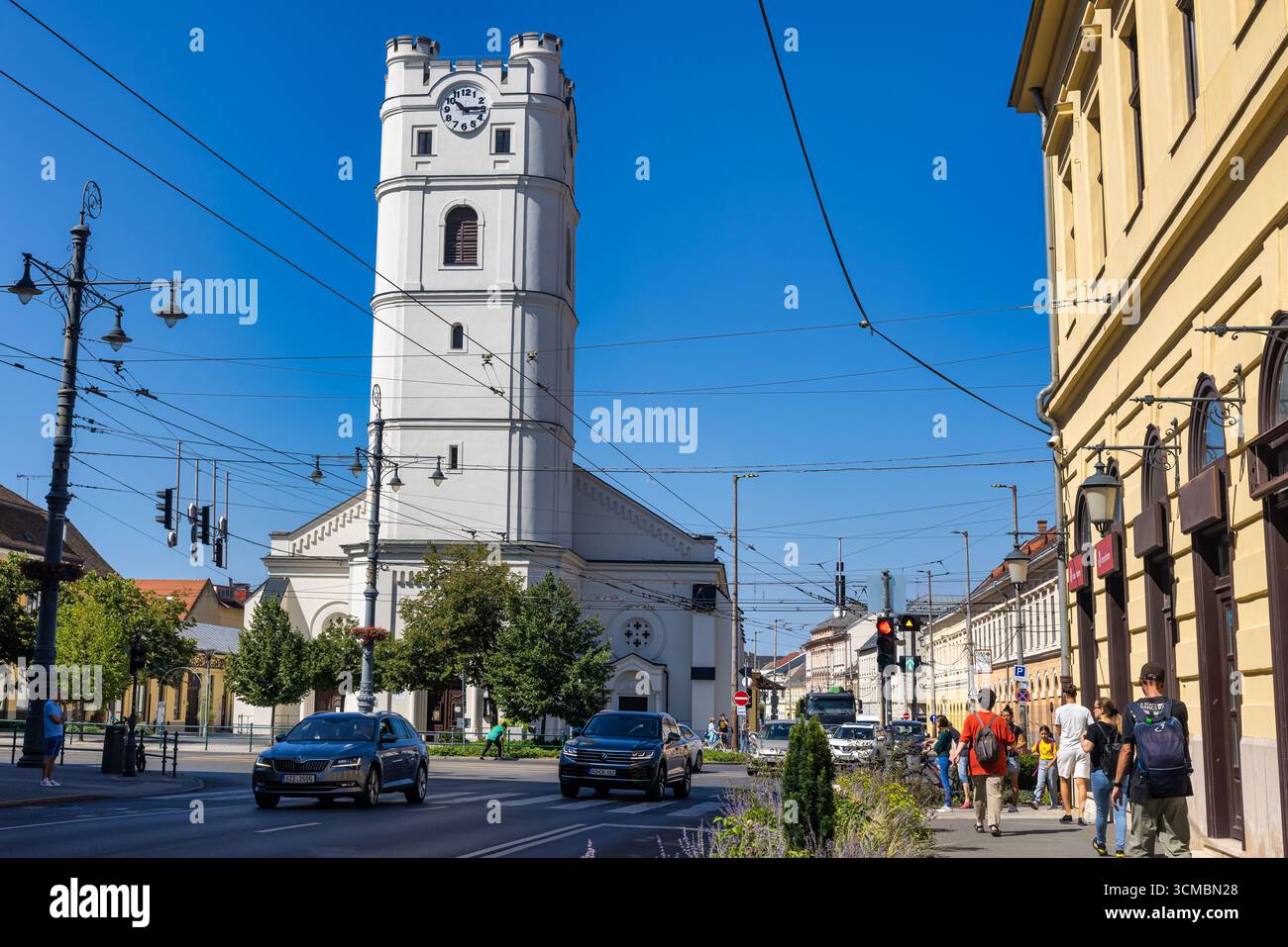 Debrecen, Ungarn 30. Juli 2025: Weißer Uhrenturm dominiert die Straßenszene im Stadtzentrum mit Fußgängern, Autos und Straßenbahnlinien unter klarem blauem Himmel. Stockfoto