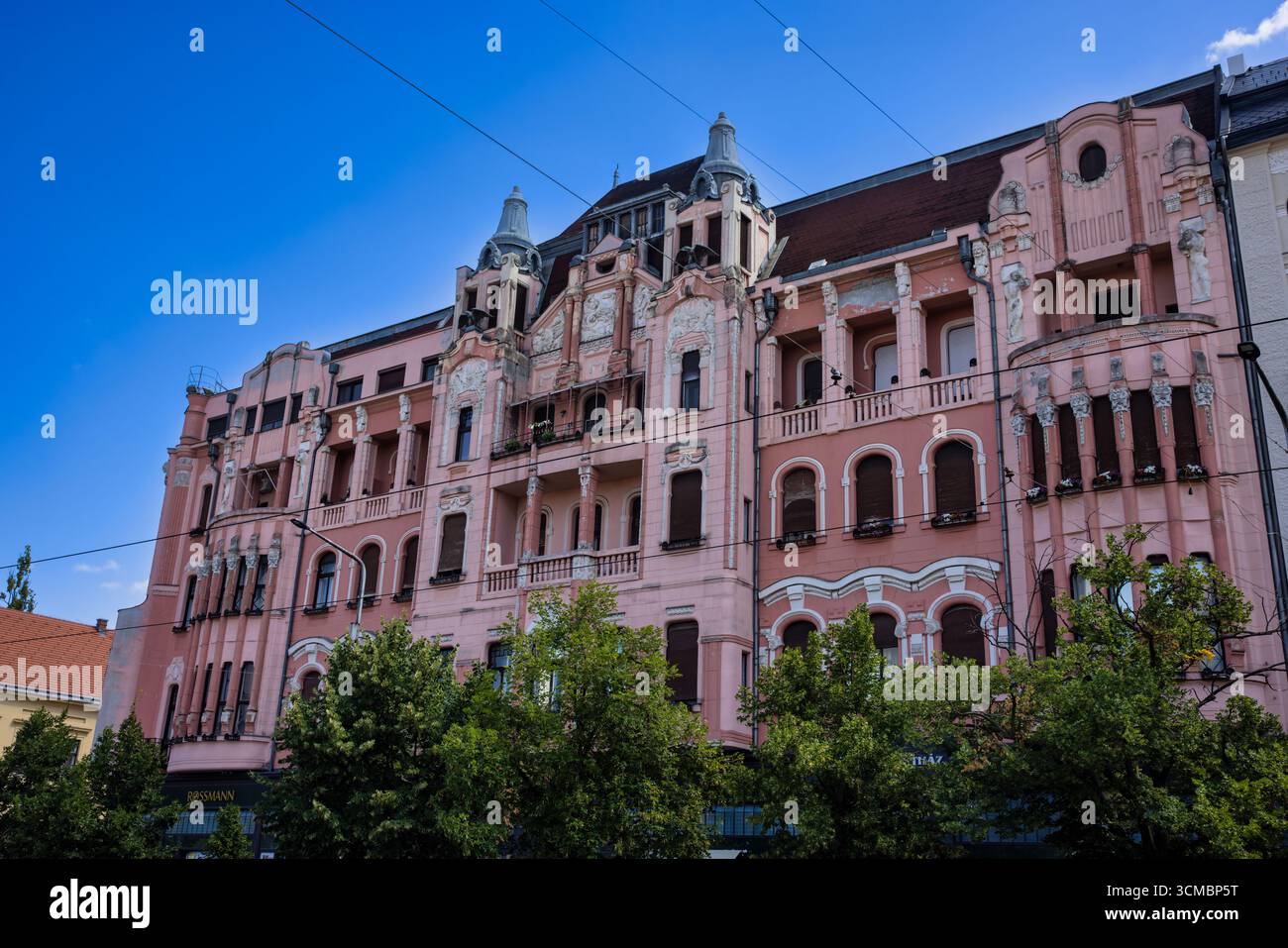Debrecen, Ungarn 30. Juli 2025 – Rosa Fassade mit Bogenfenstern, schmiedeeisernen Balkonen und Doppeltürmen im fließenden Jugendstilstil, eingerahmt von gre Stockfoto