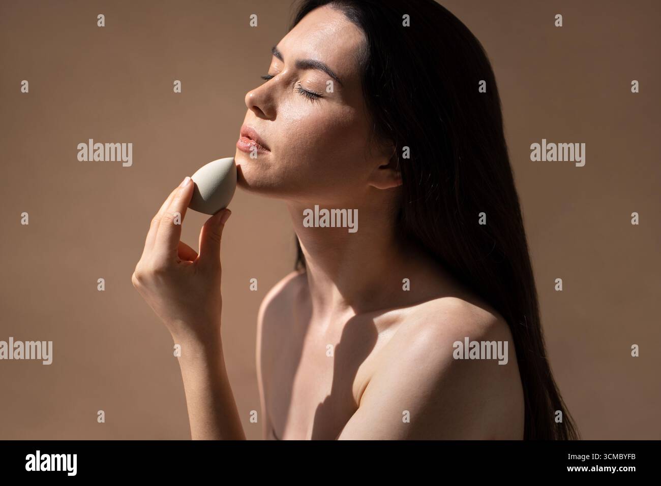 Frau schminkt sich mit einem Schönheitsschwamm und hebt ihre Hautpflegeroutine in einer ruhigen, minimalistischen Umgebung mit sanfter Beleuchtung und neutralem Ton hervor Stockfoto