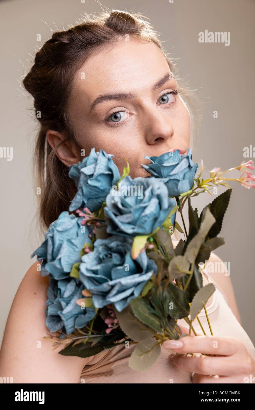 Selbstbewusste Person mit weicher Haut und natürlichem Make-up-Look hält einen Strauß blauer Rosen. Frischer strahlender Teint und anmutiger Ausdruck stehen für Selbstpflege und Selbstakzeptanz, Wellness. Stockfoto