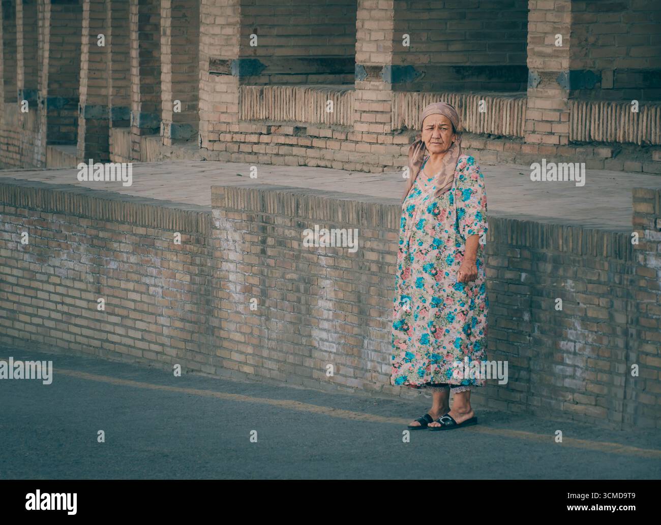Eine ältere usbekische Frau steht in Buchara, Usbekistan, an einer Backsteinmauer, in einem Blumenkleid und einem Kopftuch mit traditioneller Kleidung Stockfoto