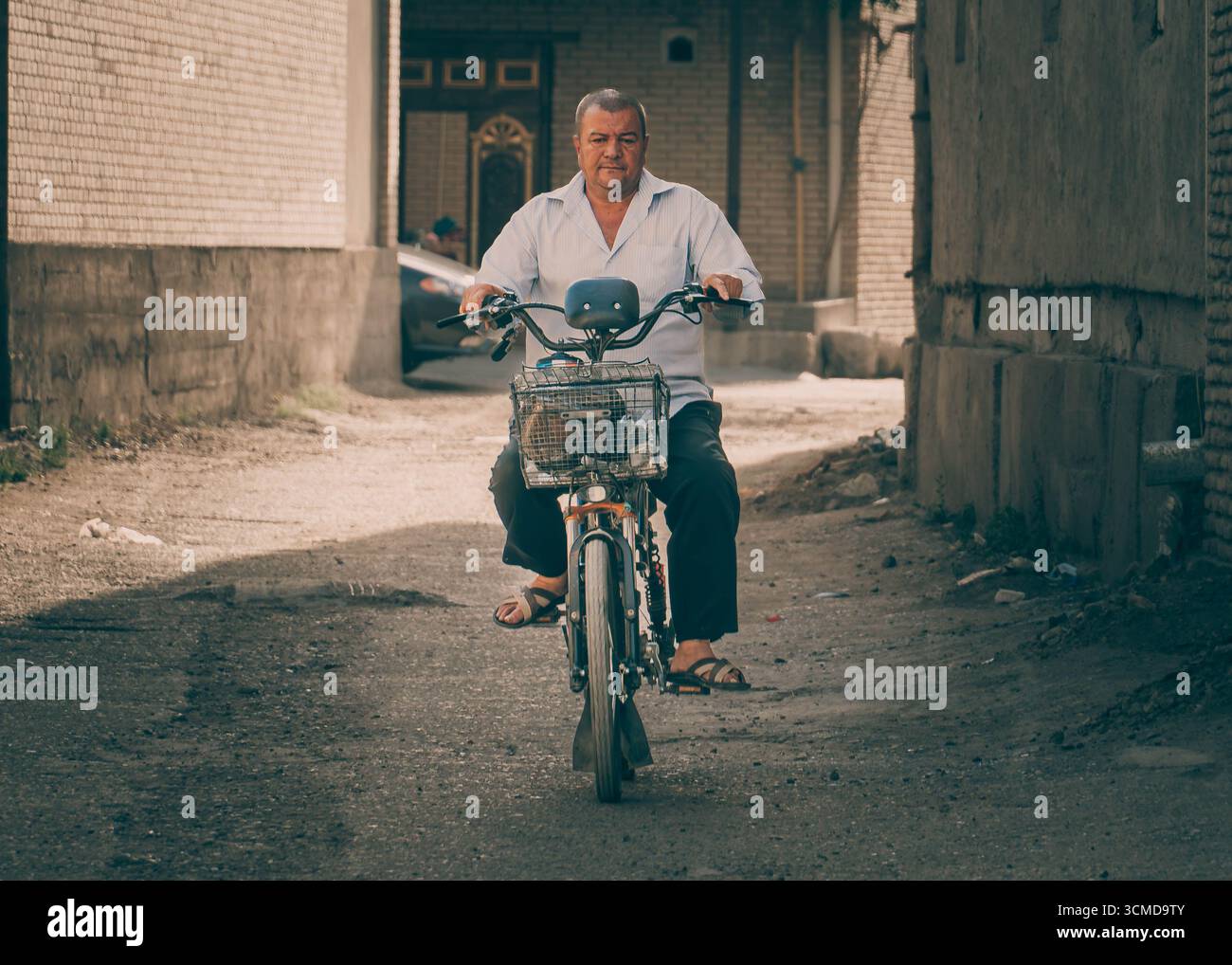 Der Usbekische Mann im mittleren Alter fährt mit dem Fahrrad durch eine verstaubte Gasse in Buchara, Usbekistan, mit Korb, weißem Hemd, dunkler Hose, und Sandalen. Stockfoto